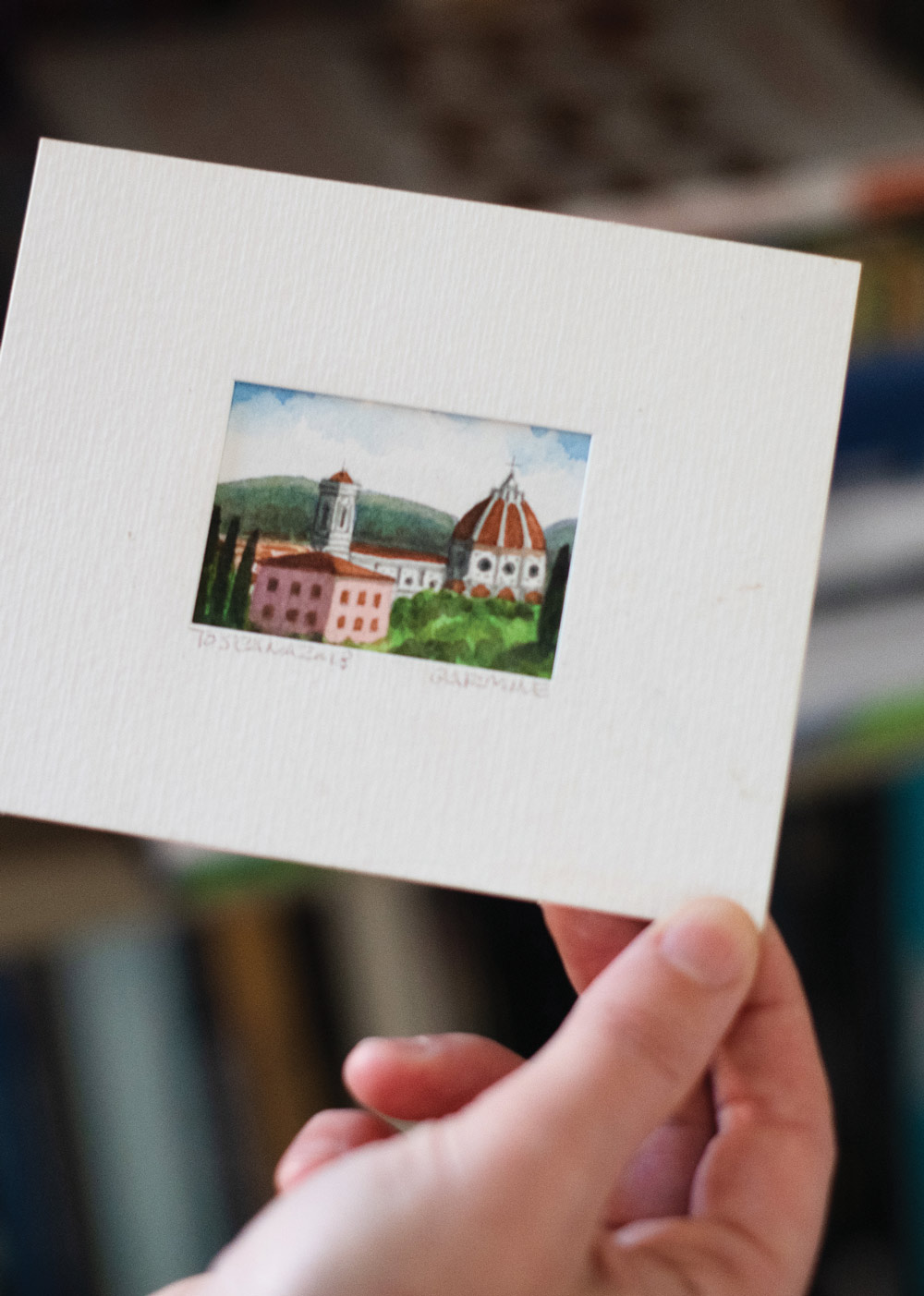 a card with a small painting of an Italian city in the center is held in hand