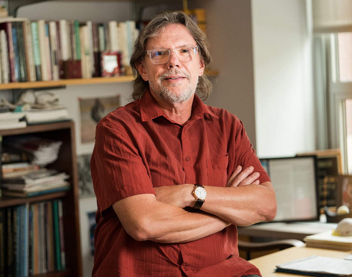 Penn GSE professor Michael Nakkula sits with his arms folded wearing a red textured button down short sleeve shirt, pictured against the background of an office