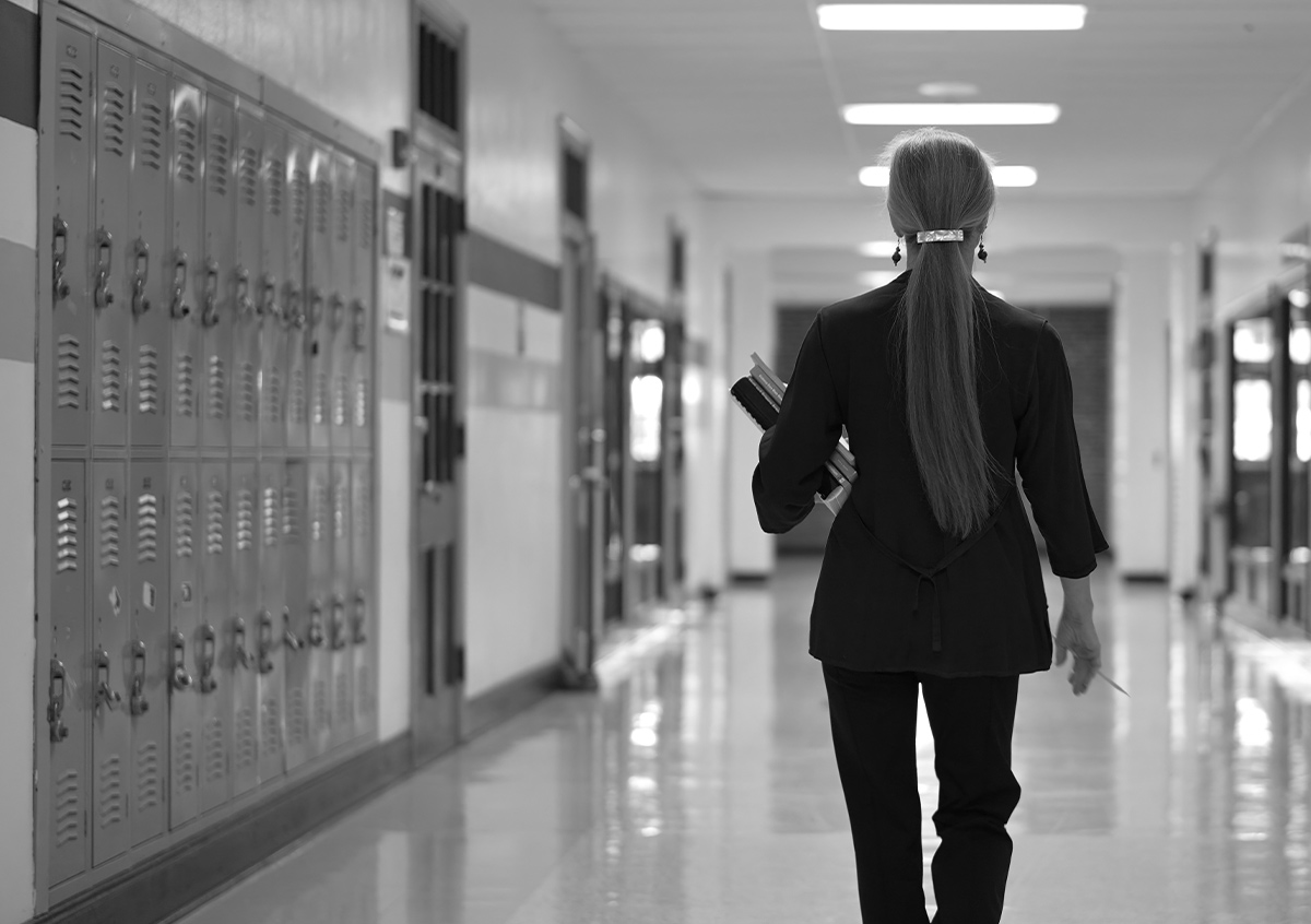 black and white image, back view of a woman walking down a school hallway lined with lockers, the woman cradles books in her left arm