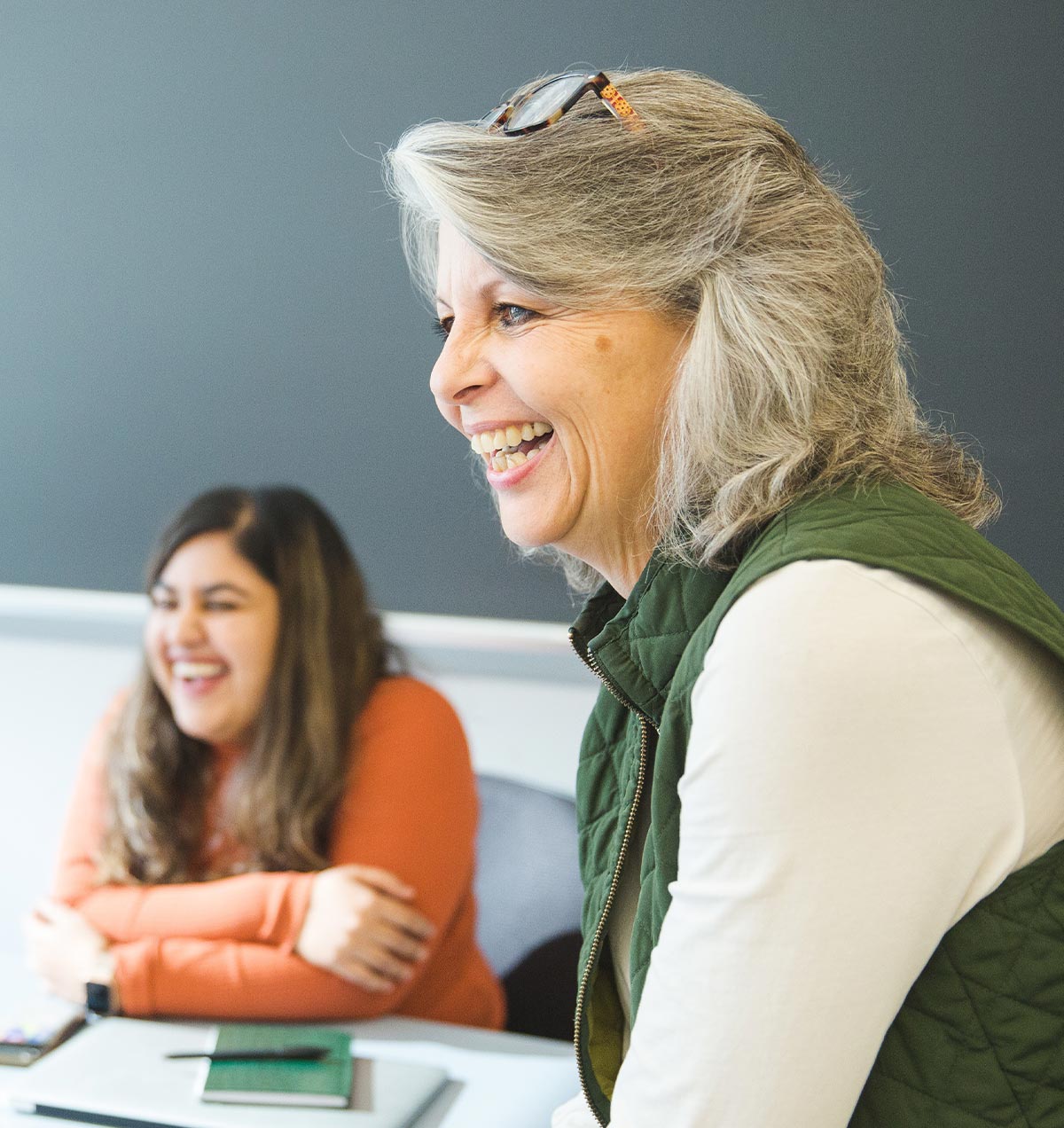 Director of School and Community Engagement Caroline Watts smiles looking off camera, out of focus behind her a student sits at a desk also smiling