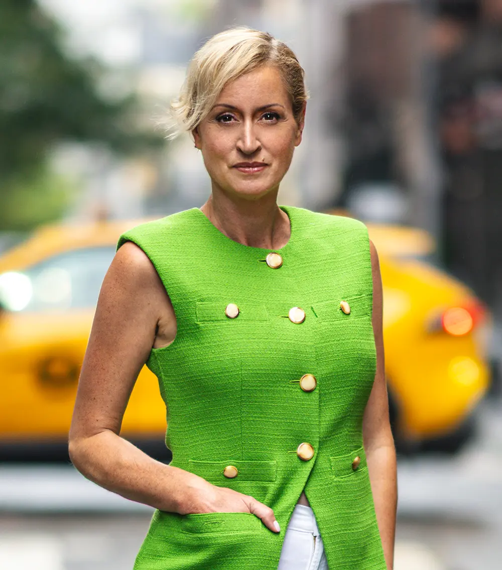 portrait image of Kandi Wiens standing in front of an out of focus, busy Pennsylvania street, she wears a serious and determined expression with her right hand in a pocket of her lime green gold buttoned blouse