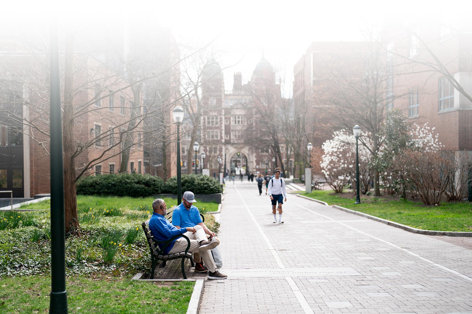 two men sitting on bench outdoors surround by trees and buildings
