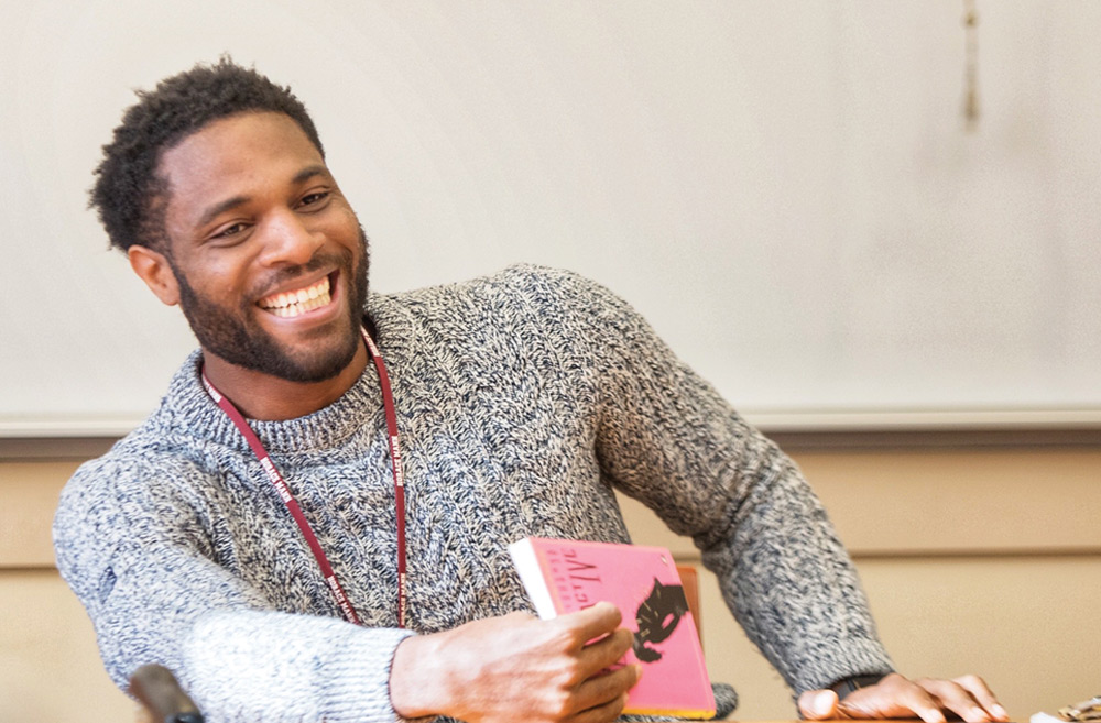 A person smiling and holding a pink book in a classroom setting, wearing a gray knit sweater.