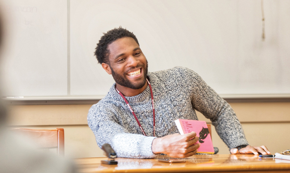 A person smiling and holding a pink book in a classroom setting, wearing a gray knit sweater.