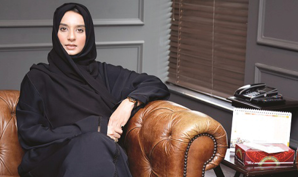 A woman in a black hijab and abaya sits on a brown leather sofa next to a telephone and calendar.